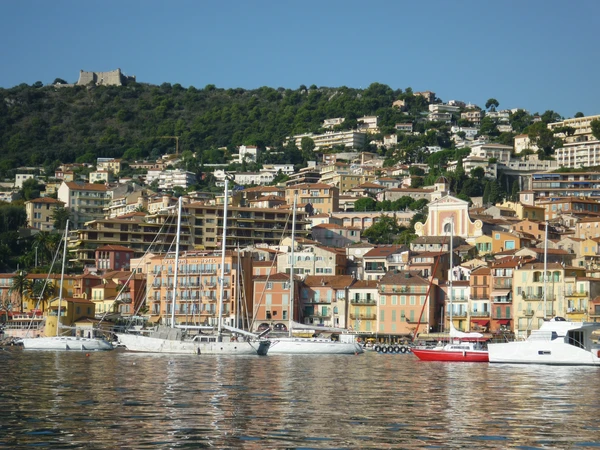 Vue panoramique de la côte méditerranéenne de Villefranche-sur-Mer en Provence-Alpes-Côte d'Azur, montrant le port naturel, les voiliers ancrés dans la baie, les bâtiments colorés du front de mer et les collines verdoyantes en arrière-plan. Photo réaliste de jour avec une lumière naturelle claire, style photographie professionnelle de destination touristique.