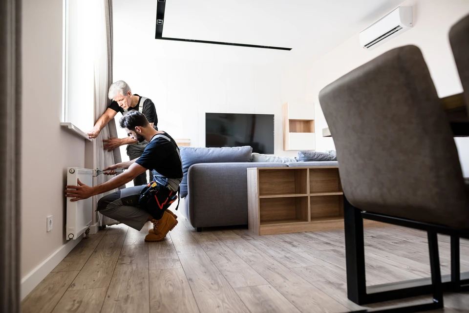 Technicien en salopette bleue effectuant la maintenance d'un radiateur à ailettes dans un intérieur résidentiel moderne